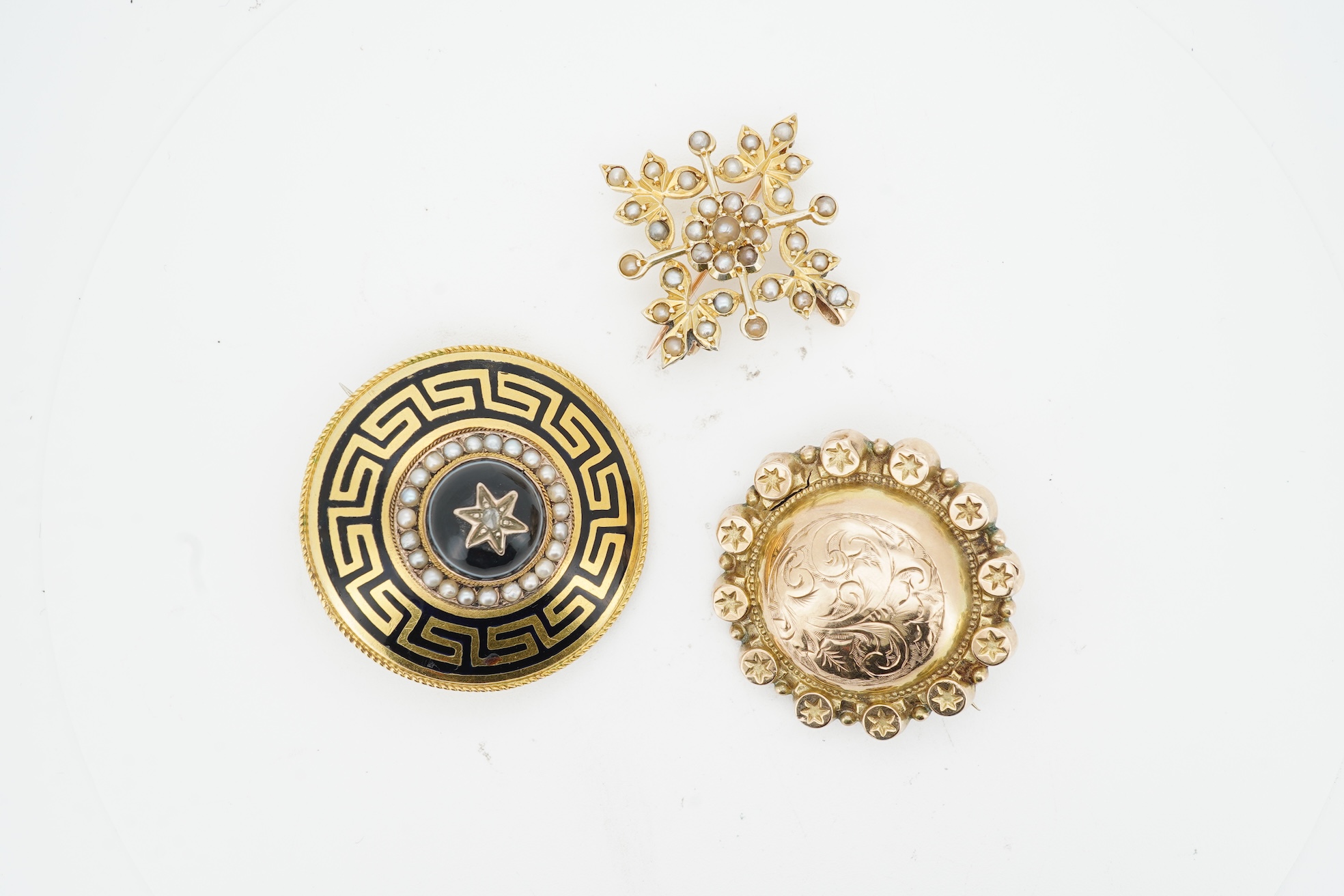 Three brooches, comprising: a Victorian circular brooch centring on a cabochon onyx set with a rose-cut diamond in a star setting, to concentring bands of seed pearls and Greek key motifs in black enamel, the reverse wit